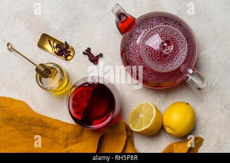 Transparency glass of hibiscus tea (karkade, Agua de flor de Jamaica) with lemon, glass teapot, honey with honey stick and orange napkin at light back Stock Photo