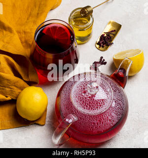 Transparency glass of hibiscus tea (karkade, Agua de flor de Jamaica) with lemon, glass teapot, honey with honey stick and orange napkin at light back Stock Photo
