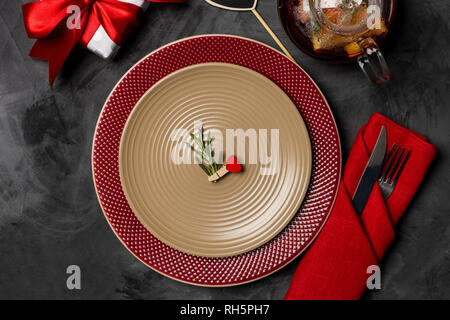 Festive table setting for Valentine's Day with fork, knife, teapot, gift box and heart pit with rosemary brunch at plate on a black table. Top view. C Stock Photo