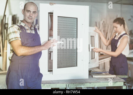 Positive workman in blue overalls demonstrating pvc window in workshop Stock Photo