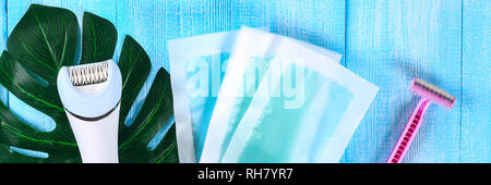 Means for hair removal. Epilator, razor for shaving, wax strips on a tropical leaf on a blue wooden background. Epilation, depilation, shaving Stock Photo