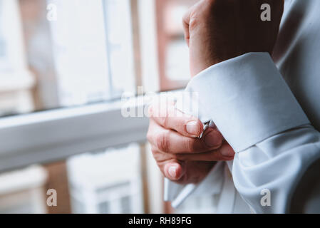 Side view of crop hands of male buttoning up white shirt sleeve near window Stock Photo