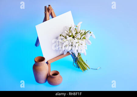 Bouquet of snowdrops and a small easel with a white paper and mini jars on a blue background. Greeting card with a space for text Stock Photo