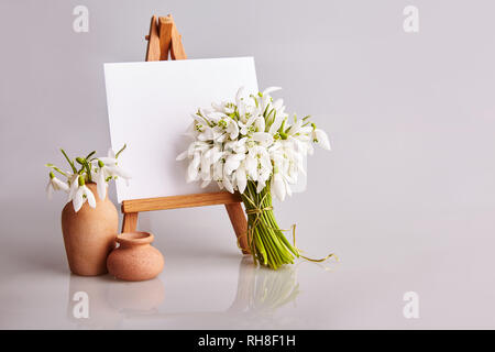 Bouquet of snowdrops and a small easel with a white paper and mini jars on a grey background. Greeting card with a space for text Stock Photo