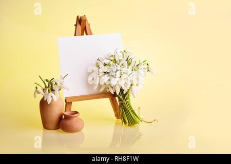 Bouquet of snowdrops and a small easel with a white paper and mini jars on a yellow background. Greeting card with a space for text Stock Photo