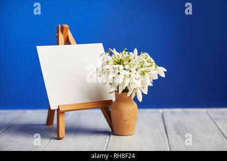 Bouquet of snowdrops and a small easel with a white paper and mini jars on a blue background. Greeting card with a space for text Stock Photo