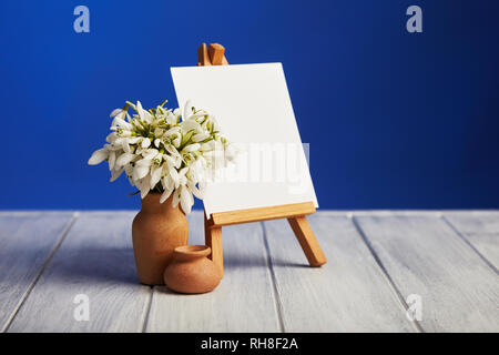 Bouquet of snowdrops and a small easel with a white paper and mini jars on a blue background. Greeting card with a space for text Stock Photo