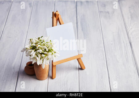 Bouquet of snowdrops and a small easel with a white paper and mini jars on a blue background. Greeting card with a space for text Stock Photo