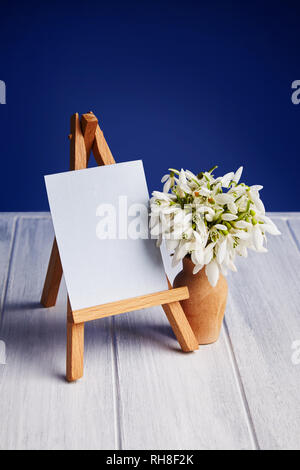 Bouquet of snowdrops and a small easel with a white paper and mini jars on a blue background. Greeting card with a space for text Stock Photo