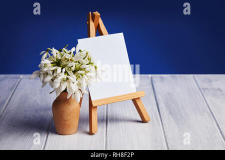Bouquet of snowdrops and a small easel with a white paper and mini jars on a blue background. Greeting card with a space for text Stock Photo