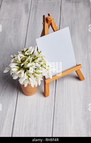 Bouquet of snowdrops and a small easel with a white paper and mini jars on a blue background. Greeting card with a space for text Stock Photo