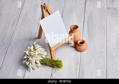 Bouquet of snowdrops and a small easel with a white paper and mini jars on a grey background. Greeting card with a space for text Stock Photo