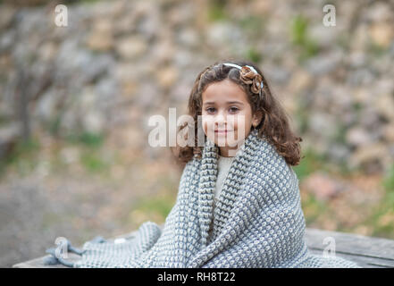 beautiful girl is sitting on nature and is warming herself with a warm knitted blanket. Stock Photo