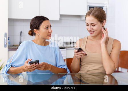 Two young women talking and using smartphone in kitchen interior Stock Photo