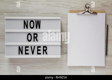 'Now or never' word on lightbox, clipboard with sheet of paper on a white wooden surface, top view. Space for text. Stock Photo