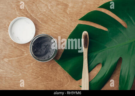 Natural toothpaste activated charcoal in glass jar and bamboo toothbrush on wooden background with green monstera leaf. Plastic free essentials, teeth Stock Photo