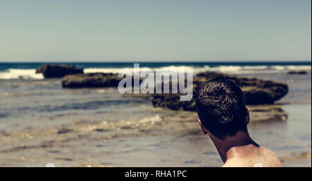 Young adult man sitting on the shore looking at the sea and the rocks with the blue sky in background and horizon Stock Photo
