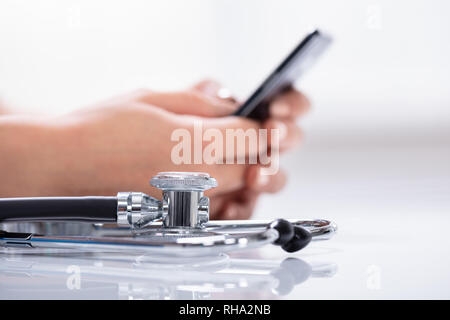 Close-up Of Stethoscope In Front Of Woman's Hand Holding Cellphone Over Reflective White Desk Stock Photo