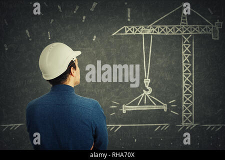 Rear view of confident young man engineer wearing white protective helmet thinking in front of blackboard. Chalk drawing and solving engineering probl Stock Photo