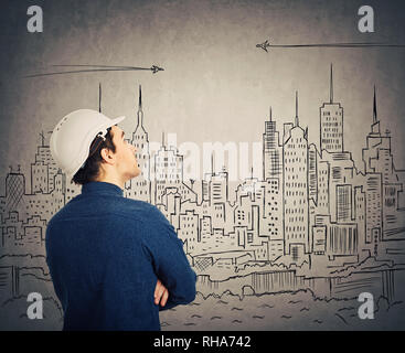 Confident young man engineer wearing protective helmet looking up and thinking of new ideas over an imaginary drawn city background. Stock Photo