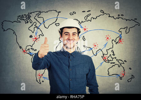 Happy cheerful young man engineer wearing protective helmet smiling showing thumb up positive gesture looking to camera isolated over world map backgr Stock Photo