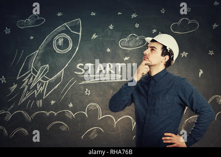 Serious thoughtful young man scientist engineer wearing protective helmet holding hand under chin pensive looking up isolated over a blackboard with r Stock Photo