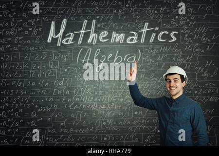 Cheerful positive thinking young man engineer wearing protective helmet showing index finger pointed up showing chalk written mathematics formulas and Stock Photo