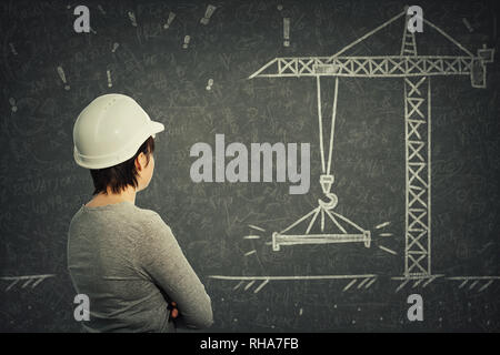 Rear view of confident young woman engineer wearing white protective helmet thinking in front of blackboard. Chalk drawing and solving engineering pro Stock Photo