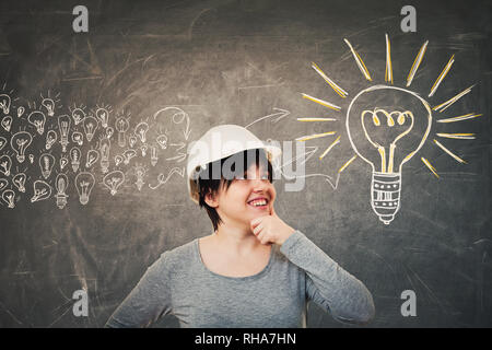 Ingenious young woman engineer wearing protective helmet holding hand under chin looking up thinking. Concept of gathering ideas into a big lightbulb. Stock Photo