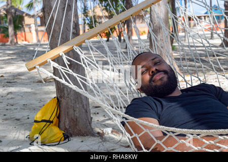 Black male mid 40s lying in hammock on tropical beach wearing black shirt looking at camera Stock Photo