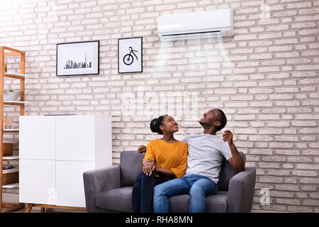 Young African Couple Sitting On Sofa Operating Air Conditioner At Home Stock Photo