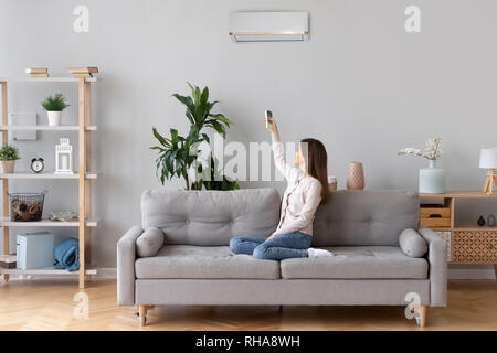 Young woman switching on air conditioner sitting on couch Stock Photo