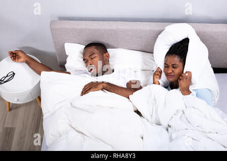 Disturbed African Woman Covering Her Ears With Pillow While Her Husband Snoring On Bed Stock Photo