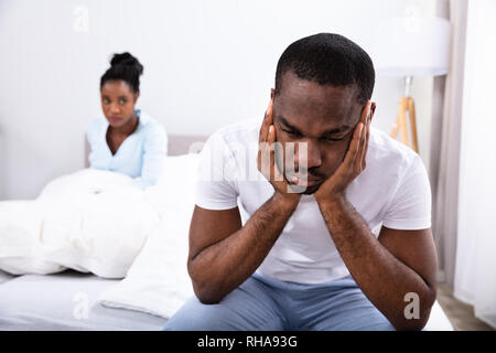 Upset Young African Man Sitting On Bed In Front Of His Wife Stock Photo