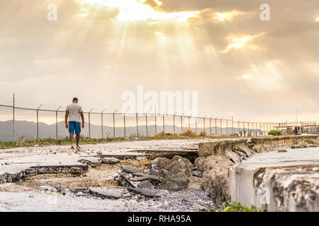 Young black male walking along road damaged by the sea, towards sun rays beaming through clouds. Concept of Hope, Broken Road, Perseverance Stock Photo