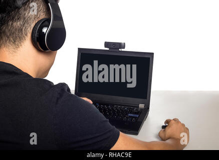 Asian male wearing headphones playing a video game and streaming online with a webcam on a laptop pc.  The image depicts entertainment industry and el Stock Photo