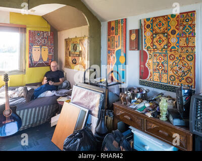 Male artist painter musician relaxing on bed looking at mobile phone in  studio bedsit sun light through window with stacked paintings Stock Photo