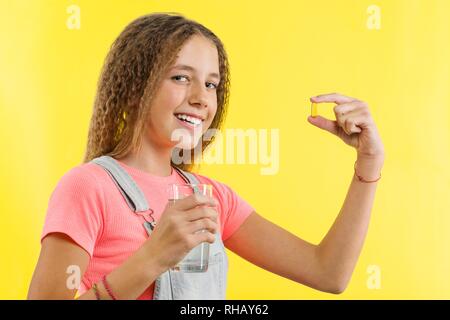 Happy smiling teen girl taking pill with cod liver oil omega-3 and holding glass of fresh water with yellow backgrounde. Diet, nutrition, healthy eati Stock Photo