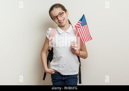 School child with a backpack holds the US flag, background bright wall in the school. Stock Photo