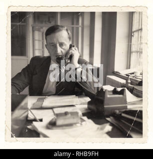 Photo of stressed unhappy older middle aged man, management, manager, managing / working in office using telephone circa 1950's, U.K. Stock Photo