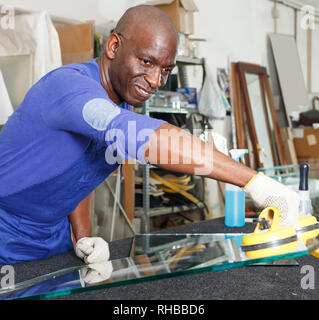 attentive African American glazier in blue overalls choosing glass for cutting in workshop Stock Photo