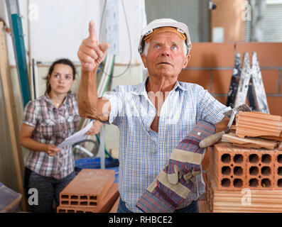 Elderly foreman in hardhat and protective gloves controlling process of construction at indoors building site Stock Photo