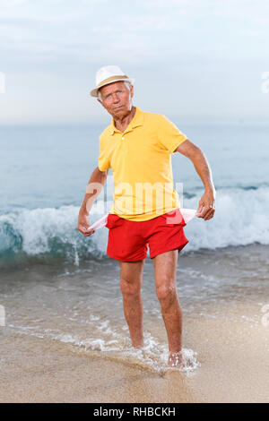 Mature man in headwear shows empty pockets near ocean Stock Photo