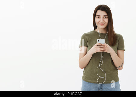 Never leave home without headphones. Portrait of tender and cute young 20s female with tattoo smiling delighted at camera holding smartphone and Stock Photo