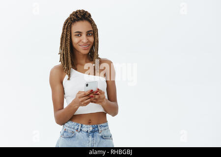 Smartphone is all you need. Portrait of stylish good-looking african american woman with cool yellow dreadlocks in white top smiling joyfully holding Stock Photo