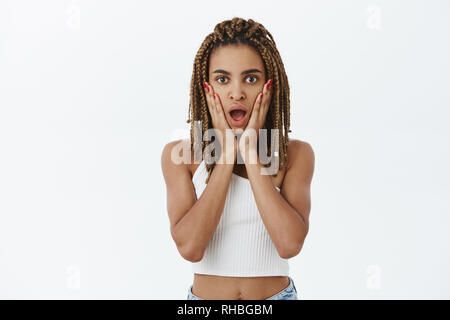Studio shot of emotive shocked and stunned young dark-skinned cool woman with yellow dreads opening mouth from amazement holding palms on cheeks Stock Photo
