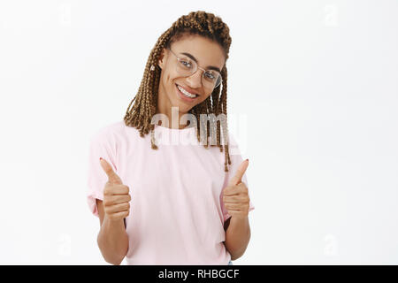 Waist-up shot of friendly and joyful gorgeous african american female with dreads in glasses and pink t-shirt showing thumbs up in like and approval Stock Photo