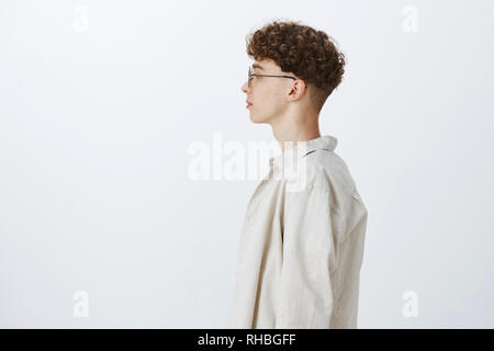Profile shot of stylish handsome young hipster male freelancer with curly hairstyle in glasses looking left and standing casually over gray background Stock Photo