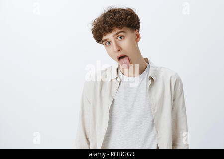 Waist-up shot of funny and handsome young male student with curly haircut in shirt showing tongue tilting head and popping eyes at camera as fooling Stock Photo