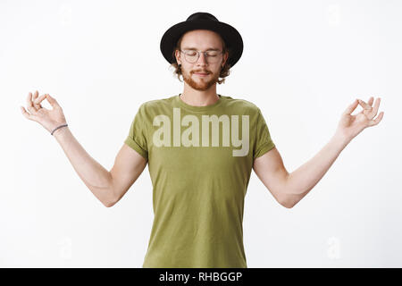 Peace in mind. Portrait of calm and relaxed attractive young bearded man with closed eyes in glasses and hat spread hands sideways with mudra orbs Stock Photo